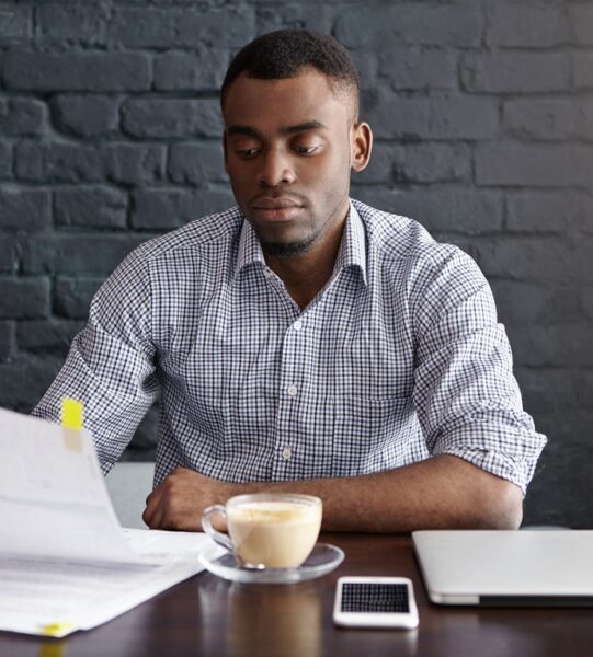 Serious attractive young businessman wearing checkered shirt reading document in his hands with attentive look while doing paperwork, analyzing finances while having lunch alone at restaurant